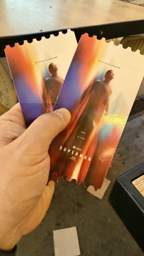 Mano sosteniendo dos entradas de cine de la nueva película de Superman, con fondo desenfocado de mesa y suelo